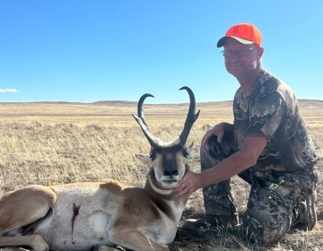 A hunter wearing a camo shirt and orange cap, kneeling beside a harvested pronghorn antelope, with Wyoming's vast grassland in the background, a proud moment captured during a guided hunt.